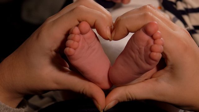 Female hands are making a heart gesture with little adorable babys feet in a center