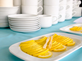 Wooden skewers lie on a stack of thin slices of lemon laid out in a stack. In the background are stacks of white cups.