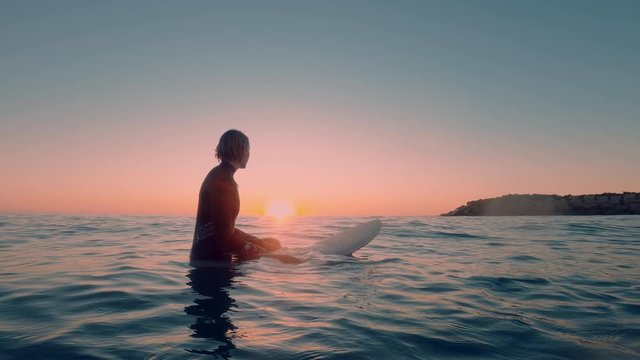 Beautiful shot from the water of a female surfer sitting on a surfboard in the blue ocean waiting for next big wave in a sunset/sunrise. Surfer easy lifestyle. Extreme sport and meditation.