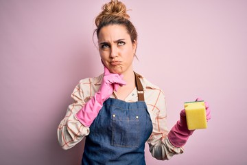 Beautiful blonde cleaner woman doing housework wearing arpon and gloves using scourer serious face...