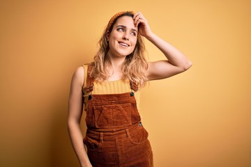Young beautiful blonde woman wearing overalls and diadem standing over yellow background smiling confident touching hair with hand up gesture, posing attractive and fashionable