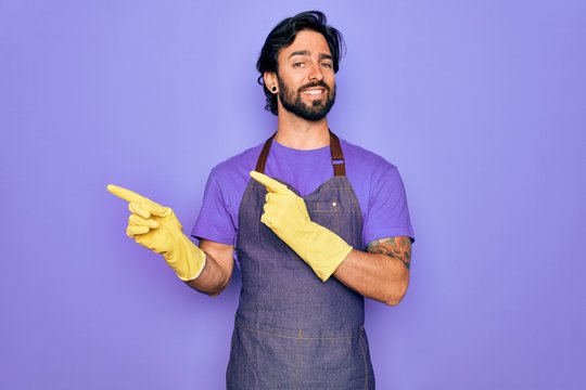 Young handsome hispanic clenaer man wearing housework apron and washing gloves smiling and looking at the camera pointing with two hands and fingers to the side.