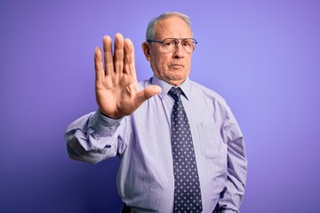 Grey haired senior business man wearing glasses standing over purple isolated background doing stop sing with palm of the hand. Warning expression with negative and serious gesture on the face.