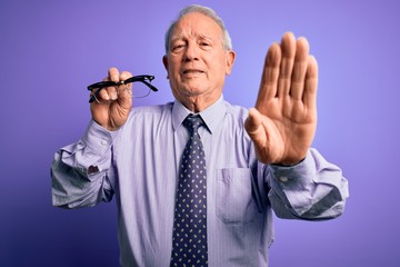 Senior grey haired man holding correction glasses over purple background with open hand doing stop sign with serious and confident expression, defense gesture