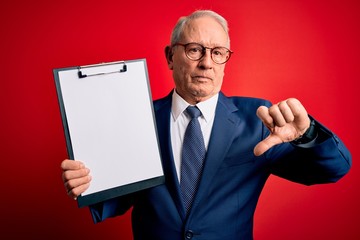 Senior grey haired business man holding clipboard over red background with angry face, negative sign showing dislike with thumbs down, rejection concept