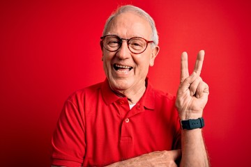 Grey haired senior man wearing glasses and casual t-shirt over red background smiling with happy face winking at the camera doing victory sign. Number two.