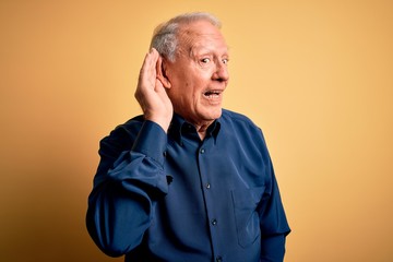 Grey haired senior man wearing casual blue shirt standing over yellow background smiling with hand over ear listening an hearing to rumor or gossip. Deafness concept.