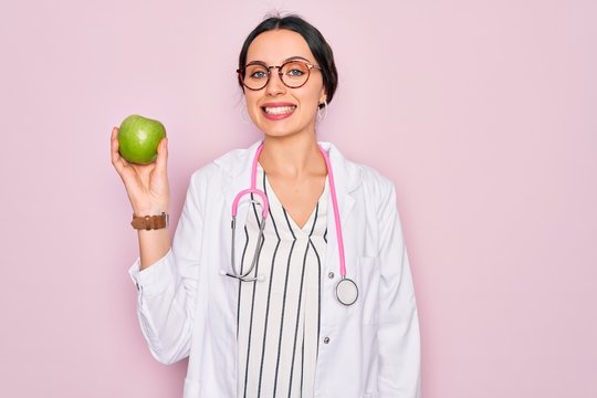 Young beautiful doctor woman with blue eyes wearing stethoscope holding green apple fruit with a happy face standing and smiling with a confident smile showing teeth