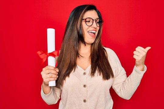 Young hispanic smart woman wearing glasses holding university degree over red background pointing and showing with thumb up to the side with happy face smiling