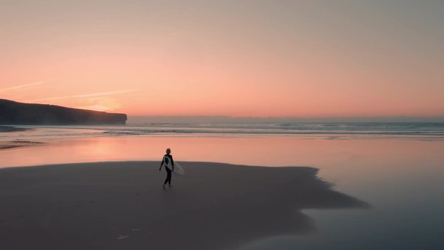 Beautiful aerial view of attractive surfer woman walking on the sand towards the ocean at sunset / sunrise carrying her surfboard. She is going out to surf.