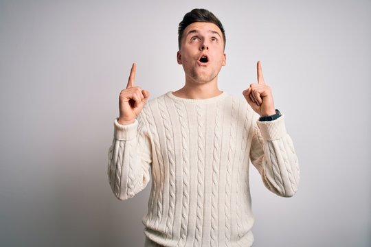 Young handsome caucasian man wearing casual winter sweater over white isolated background amazed and surprised looking up and pointing with fingers and raised arms.
