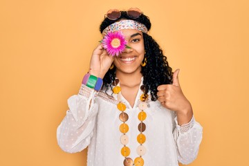 Young african american curly hippie woman wearing sunglasses holding pink flower on eye happy with big smile doing ok sign, thumb up with fingers, excellent sign
