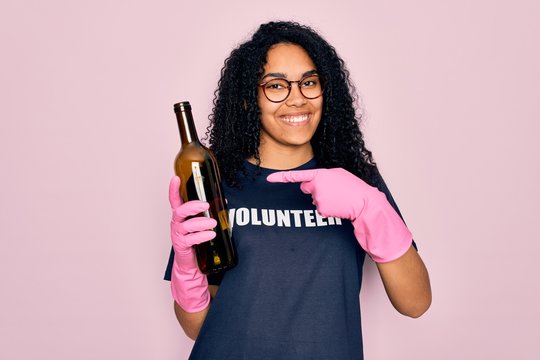 African american curly woman wearing volunteer t-shirt doing volunteering recycling glass bottle very happy pointing with hand and finger