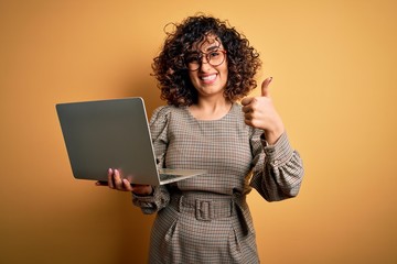 Beautiful arab businesswoman wearing glasses working using laptop over yellow background happy with big smile doing ok sign, thumb up with fingers, excellent sign