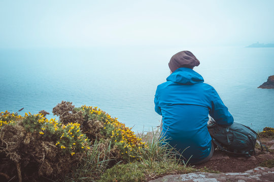 Young man with backpack, raincoat and wooly hat sitting on the edge of a coast cliff watching the sea on a rainy and moody day. Outdoors toned image with copy space.