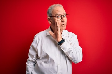 Middle age handsome hoary man wearing casual shirt and glasses over red background hand on mouth telling secret rumor, whispering malicious talk conversation