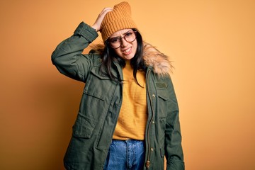 Naklejka na ściany i meble Young brunette woman wearing glasses and winter coat with hat over yellow isolated background confuse and wonder about question. Uncertain with doubt, thinking with hand on head. Pensive concept.
