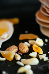 Sweet homemade stack of pancakes with honey filling, almonds and dried apricots, plate on black background. Healthy vegetarian food during quarantine