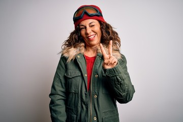 Middle age skier woman wearing snow sportswear and ski goggles over white background smiling with happy face winking at the camera doing victory sign. Number two.