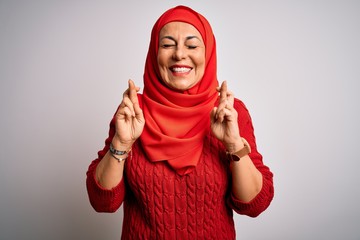 Middle age brunette woman wearing muslim traditional hijab over isolated white background gesturing finger crossed smiling with hope and eyes closed. Luck and superstitious concept.