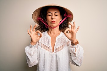 Middle age brunette woman wearing asian traditional conical hat over white background relax and smiling with eyes closed doing meditation gesture with fingers. Yoga concept.