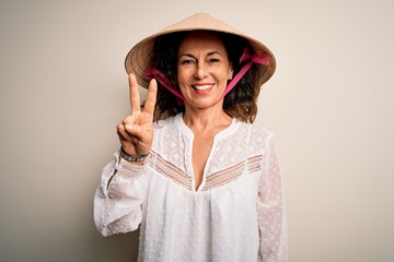 Middle age brunette woman wearing asian traditional conical hat over white background showing and pointing up with fingers number two while smiling confident and happy.