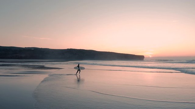 Beautiful aerial view of attractive surfer woman running on the sand towards the ocean at sunset / sunrise going to surf. Surrounded by an amazing landscape. Fun day at the beach surfing.