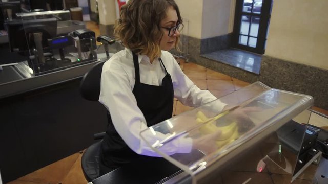 Cheerful caucasian saleswoman in white shirt and black apron scanning product, fruits at checkout counter in bright supermarket and putting it into brown paper bag, take an EC from customer and swipe