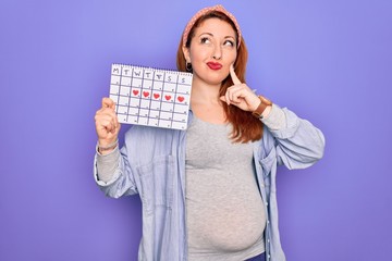 Young beautiful redhead pregnant woman expecting baby holding period calendar serious face thinking about question, very confused idea