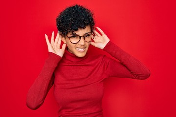 Fototapeta na wymiar Young beautiful african american afro woman wearing turtleneck sweater and glasses Trying to hear both hands on ear gesture, curious for gossip. Hearing problem, deaf