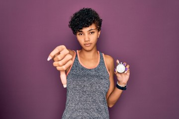 Young beautiful african american sporty woman holding stopwatch over purple background with angry...