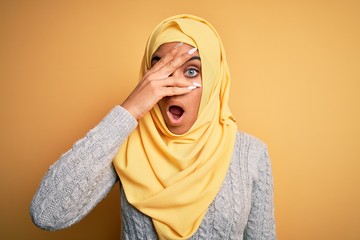 Young beautiful african american girl wearing muslim hijab over isolated yellow background peeking in shock covering face and eyes with hand, looking through fingers with embarrassed expression.