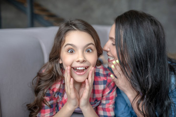 Dark-haired mom telling something interesting to her surprised daughter in the ear