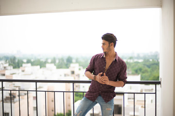 Portrait of an young and handsome brunette Bengali muscular man in casual shirt and jeans standing on a balcony in white urban background. Indian lifestyle.