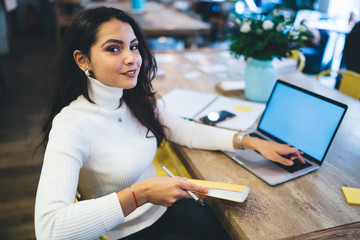 Portrait of attractive woman with education literature book in hand looking at camera during e learning time in coworking space, beautiful Caucasian millennial doing remote job on netbook technology