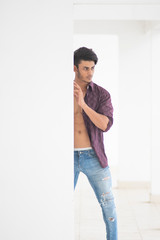 Fototapeta na wymiar An young and handsome brunette Bengali muscular in open shirt and blue jeans peeping out from a pillar in white background. Indian lifestyle.