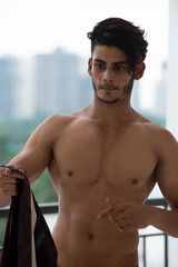 Fototapeta na wymiar Portrait of an young and handsome brunette Bengali muscular man standing in bare body on a balcony in green urban background. Indian lifestyle.