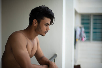 Naklejka na ściany i meble Portrait of an young and handsome brunette Bengali muscular man in bare body smoking on a balcony in white urban background. Indian lifestyle.