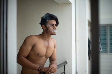 Fototapeta na wymiar Portrait of an young and handsome brunette Bengali muscular man in bare body smoking on a balcony in white urban background. Indian lifestyle.