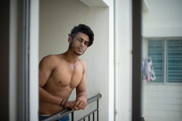 Obraz na płótnie Canvas Portrait of an young and handsome brunette Bengali muscular man in bare body smoking on a balcony in white urban background. Indian lifestyle.