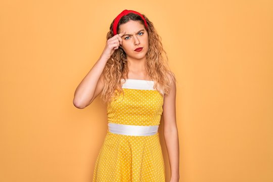 Beautiful blonde pin-up woman with blue eyes wearing diadem standing over yellow background pointing unhappy to pimple on forehead, ugly infection of blackhead. Acne and skin problem