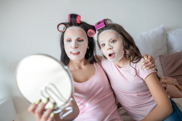 Mom and daughter with facial mask on their faces looking at themselves in the mirror