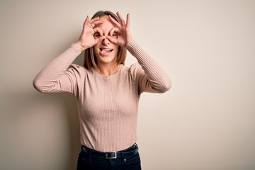 Young beautiful woman wearing casual sweater standing over isolated white background doing ok gesture like binoculars sticking tongue out, eyes looking through fingers. Crazy expression.