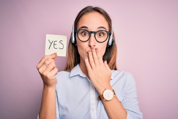 Beautiful call center agent woman working using headset holding reminder with yes message cover...
