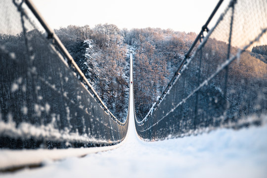 Verschneite H&auml;ngeseilbr&uuml;cke mit Person in der Mitte umgeben von verschneiten B&auml;umen im Morgengrauen mit buntem Licht. 