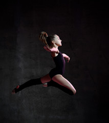 Fototapeta na wymiar Young girl gymnast in a black sports body and special tops jumps in a gymnastic pose on a dark background