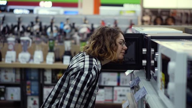 Side view of a young curly haired woman in plaid shirt chooses a microwave oven in a consumer electronics supermarket. Opens the door and looking inside the oven