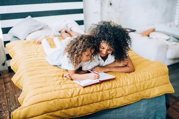 Black mother and daughter lying on bed and doing homework together