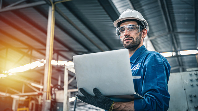 Confident engineer in blue jumpsuit holding laptop computer in a warehouse.