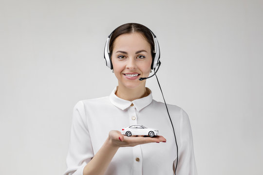 Car insurance. Beautiful smiling woman consultant of call center in headphones holding white car on gray background. female customer support operator with headset